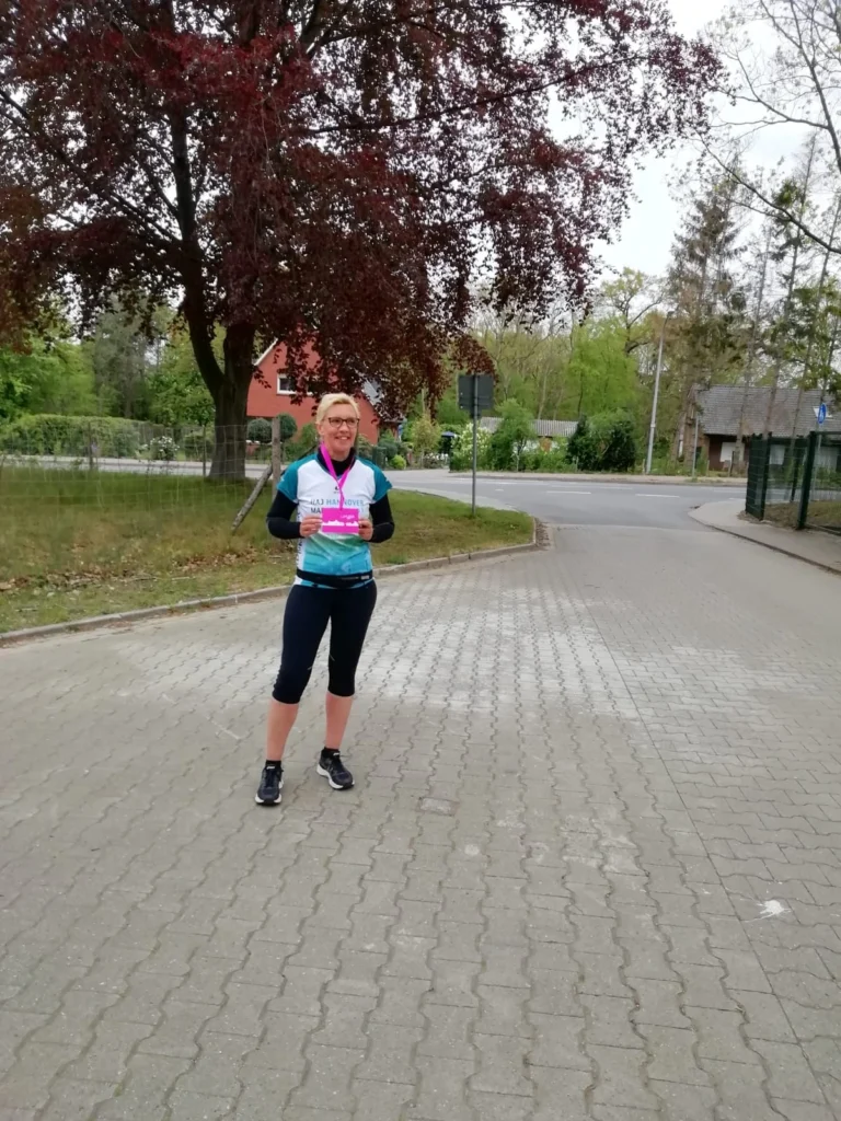 Läuferin in Sportbekleidung steht auf einer gepflasterten Straße und hält eine Medaille in der Hand. Im Hintergrund sind Bäume und ein Wohnhaus zu sehen, was eine aktive Outdoor-Umgebung vermittelt.