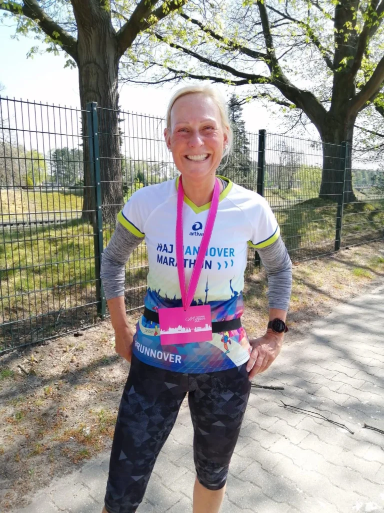 Läuferin nach dem Hannover Marathon, trägt ein sportliches Outfit mit Medaille und Teilnehmerausweis, steht lächelnd im Freien vor einem Zaun und Bäumen.