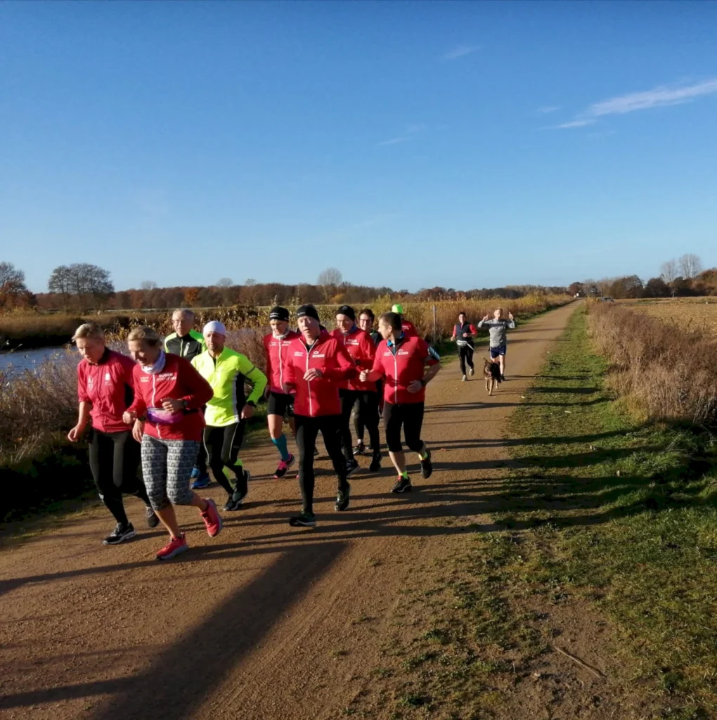 Läufergruppe in roten Jacken joggt auf einem Weg entlang eines Flusses an einem sonnigen Herbsttag, umgeben von buntem Laub und einer klaren blauen Himmel.