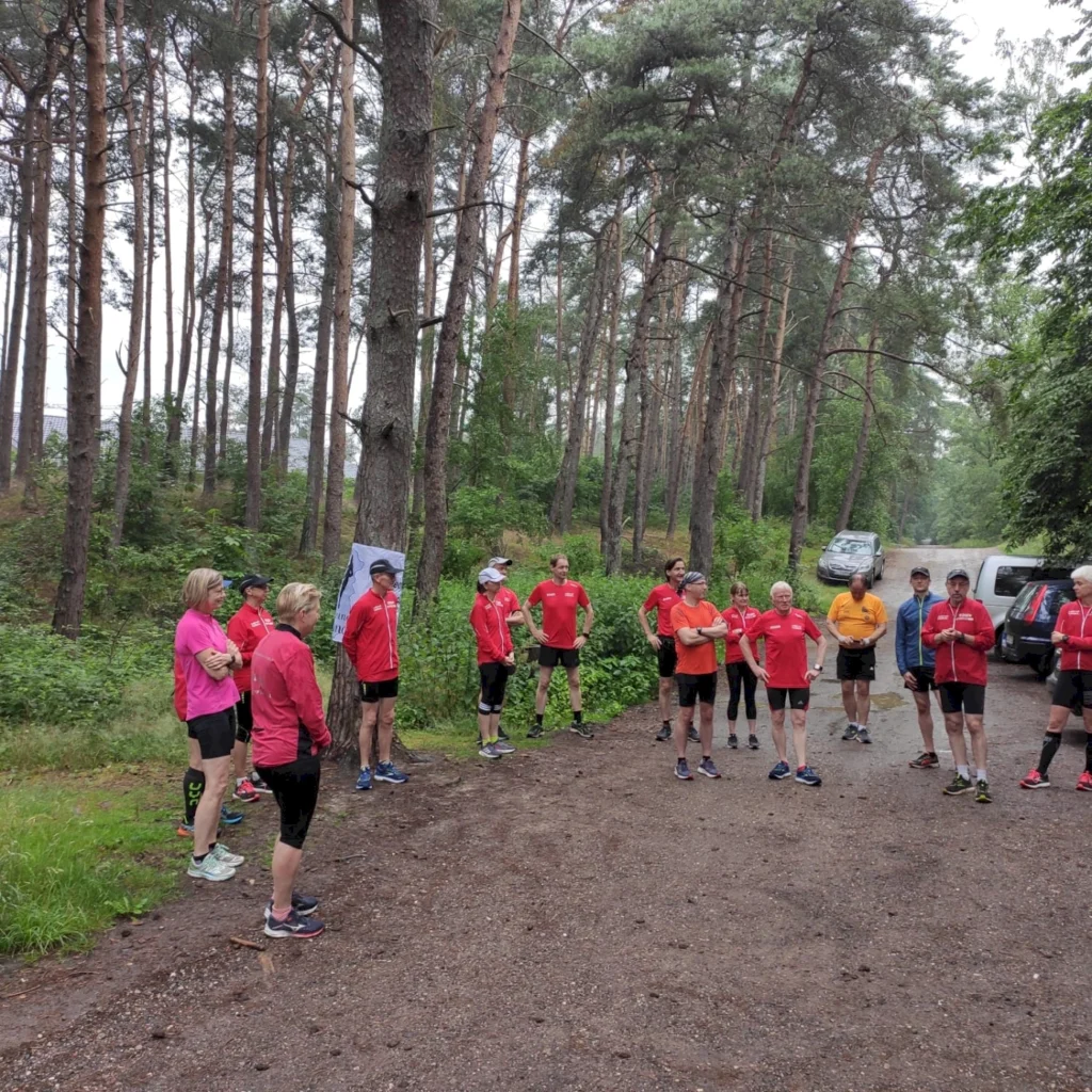 Gruppe von Läufern in roten Shirts versammelt sich im Wald, um sich auf ein Training oder einen Wettkampf vorzubereiten. Umgeben von hohen Bäumen und grüner Vegetation, herrscht eine motivierende Atmosphäre.