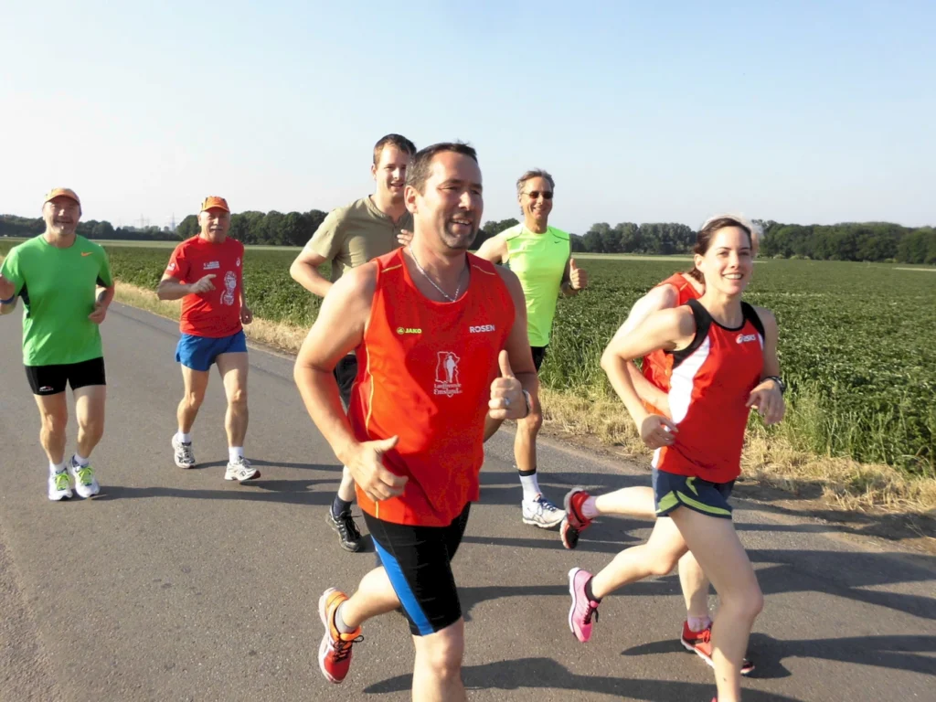 Gruppe von Läufern in sportlicher Kleidung, die auf einer Landstraße joggen, umgeben von grünen Feldern und klarem Himmel.