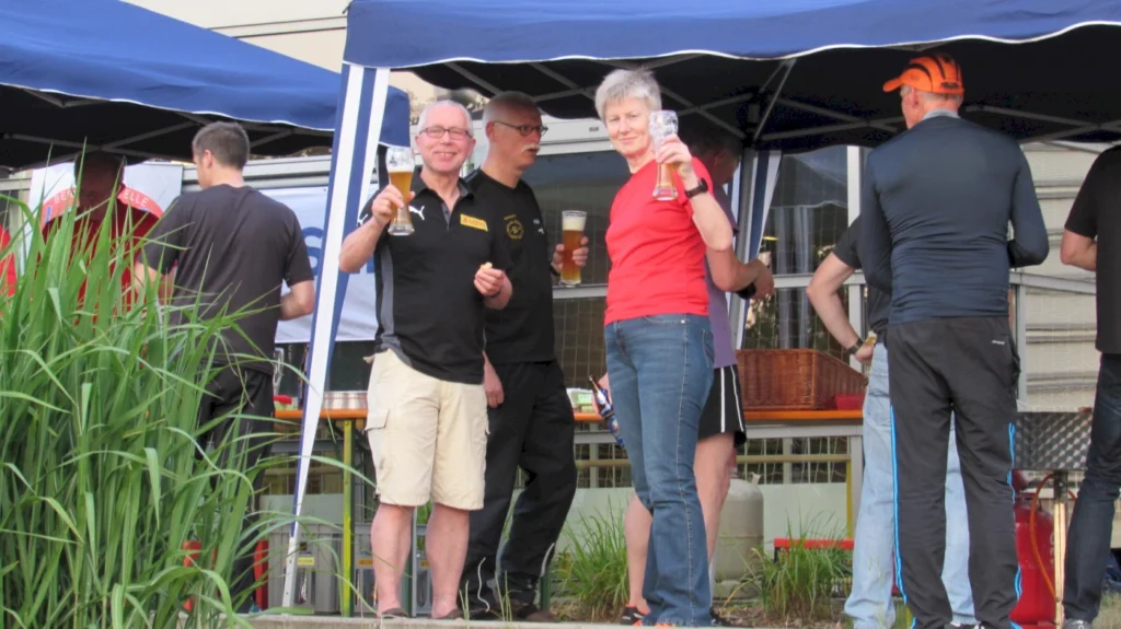 Menschen genießen Bier unter Pavillon auf Sommerfest im Freien. Garten und Zelte im Hintergrund. Feierliche Stimmung.