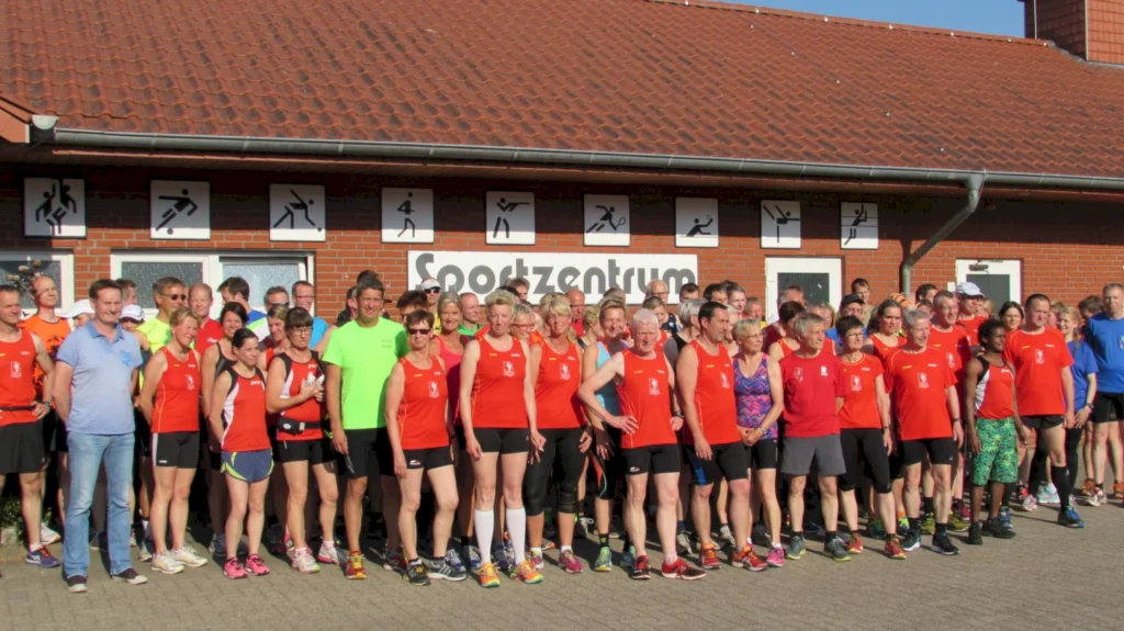 Gruppe von Läufern in roten Trikots vor einem Sportzentrum bei Sonnenschein, bereit für ein Rennen oder Training.