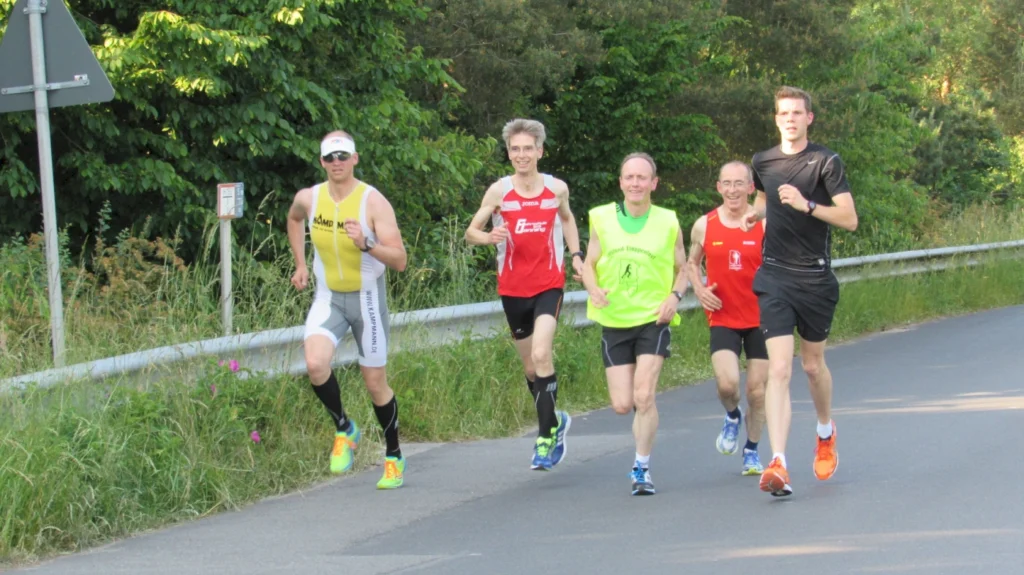 Fünf Männer joggen gemeinsam auf einer Straße im Grünen bei sonnigem Wetter.