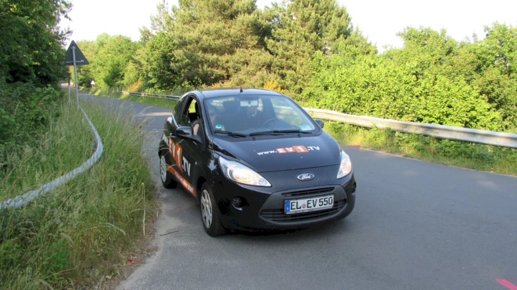 Schwarzer Ford Fiesta auf einer Landstraße, umgeben von grüner Vegetation und einem Straßenschild im Hintergrund, mit der Website www.EV1.tv auf der Seite des Fahrzeugs.