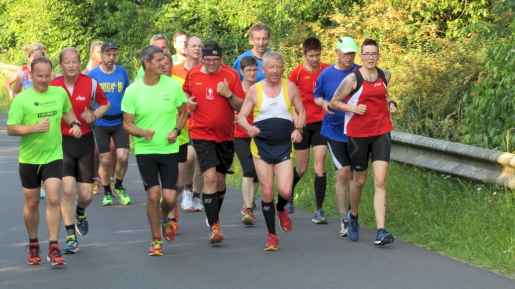 Gruppe von Läufern in bunter Sportkleidung joggt auf einer Straße im Grünen bei sonnigem Wetter.