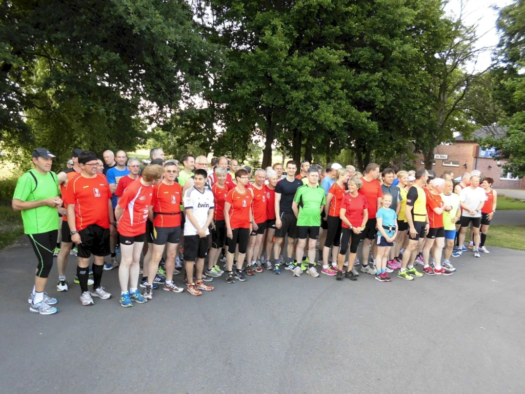 Gruppe von Läufern in bunter Sportkleidung steht auf der Straße vor einem Park, bereit zum gemeinsamen Lauf-Event.