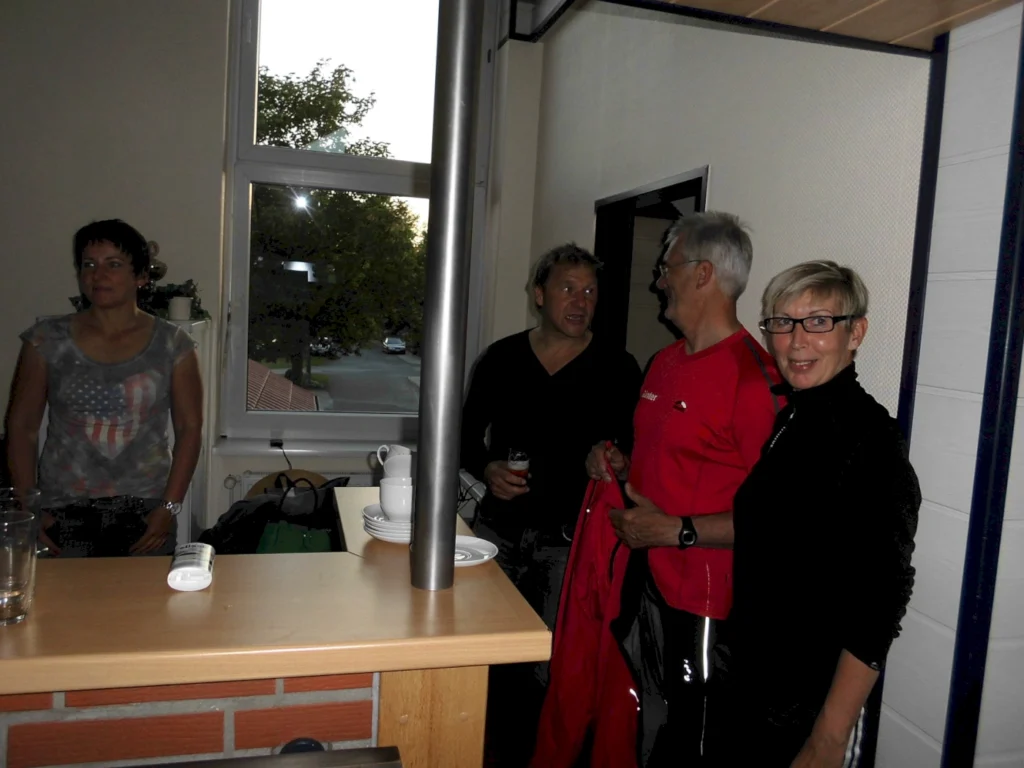 Vier Personen stehen in einem Raum mit Fenstern, während sie sich unterhalten. Eine Frau mit einem T-Shirt mit amerikanischem Motiv blickt nachdenklich, während die anderen drei Personen, darunter ein Mann in einem roten Shirt, freundlich miteinander lachen. Auf einem Tisch sind Gläser und Geschirr sichtbar.