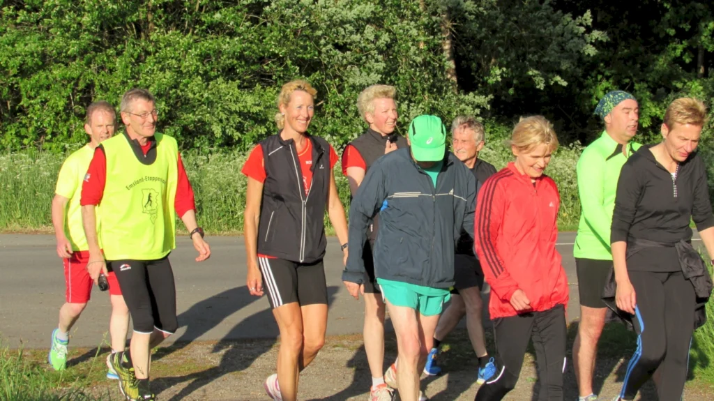 Gruppe von Personen joggt im Freien an einem sonnigen Tag auf einer Straße, umgeben von grünen Bäumen.