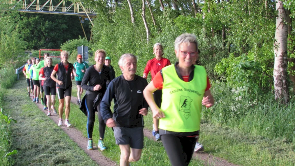 Gruppe von Läufern joggt fröhlich auf einem Waldweg, umgeben von Bäumen und frischem Grün.