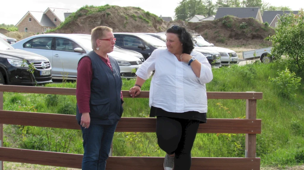 Zwei Frauen unterhalten sich draußen vor parkenden Autos, im Hintergrund sind Häuser zu sehen.