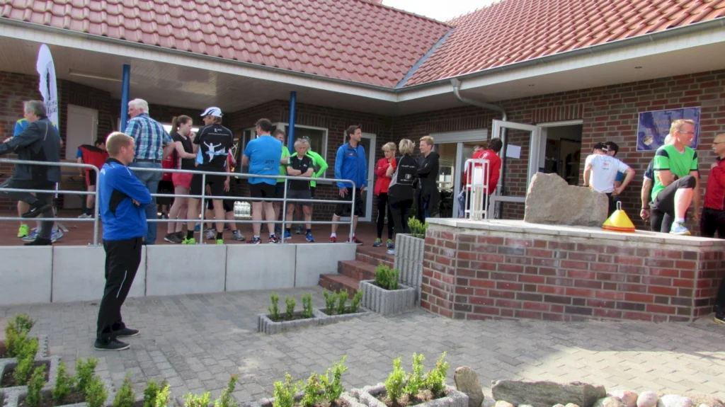 Gruppierung von Laufbegeisterten vor einem Sportgebäude, die sich auf eine Veranstaltung vorbereiten. Einige Teilnehmer tragen Sportbekleidung in verschiedenen Farben, während andere in Gespräche vertieft sind. Im Hintergrund sind Wasserflaschen und ein Banner sichtbar, das auf die Veranstaltung hinweist.