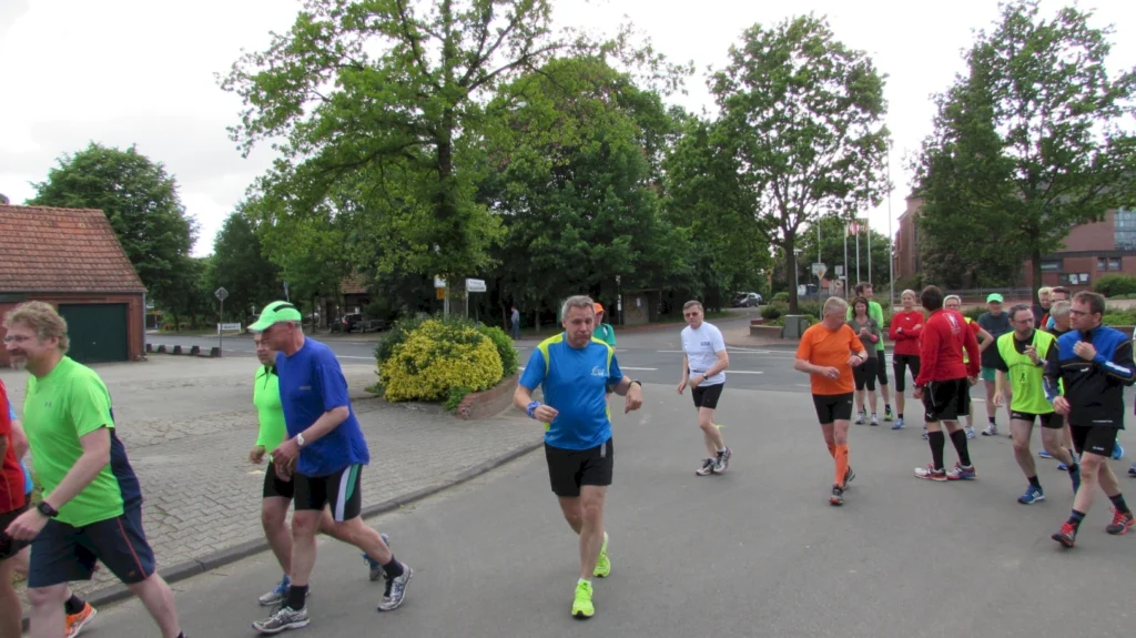Gruppe von Läufern in bunter Sportkleidung bei einem Stadtlauf, von Bäumen umgeben, an einer Kreuzung.
