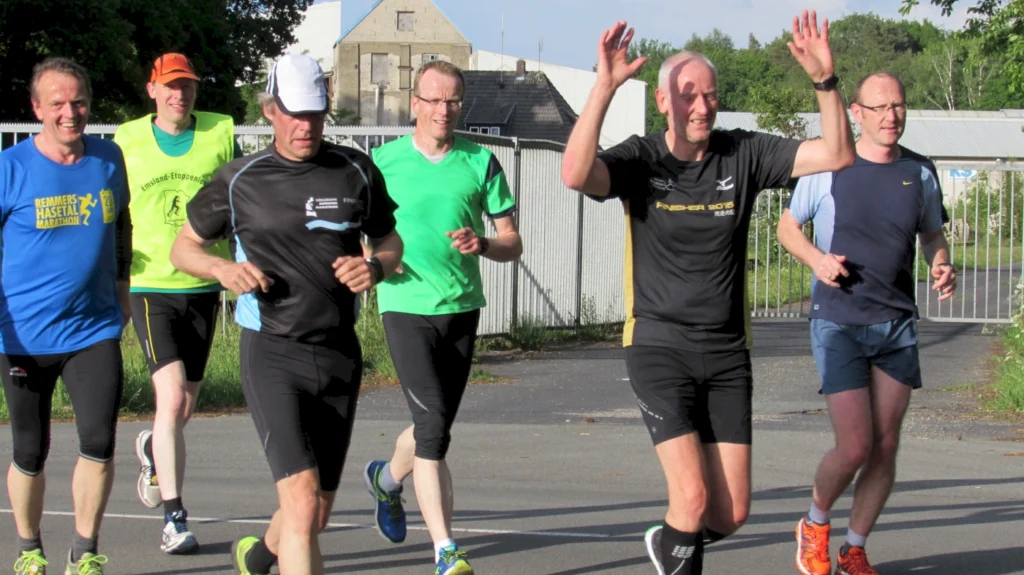 Gruppe von fünf Männern in Sportkleidung, die gemeinsam joggen, lächelnd und motiviert, auf einem asphaltierten Weg im Freien, umgeben von Bäumen und Gebäuden. Ideal für Themen wie Laufen, Fitness und Gemeinschaftssport.