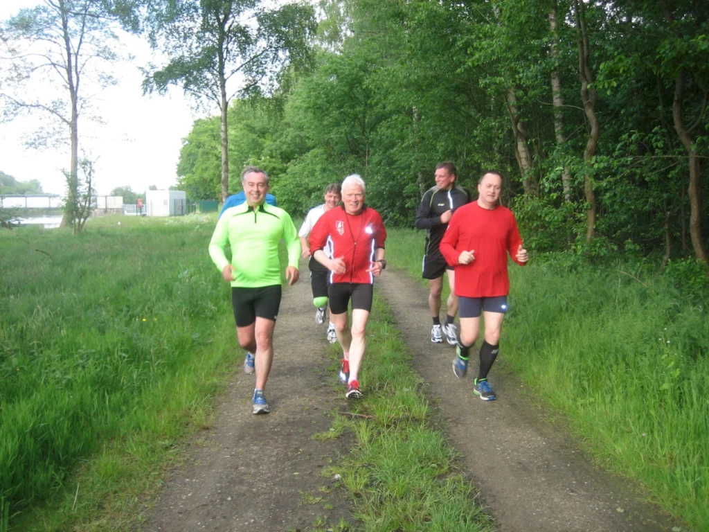 Fünf Männer joggen auf einem Waldweg, umgeben von Bäumen und grüner Natur, bei hellem Tageslicht.