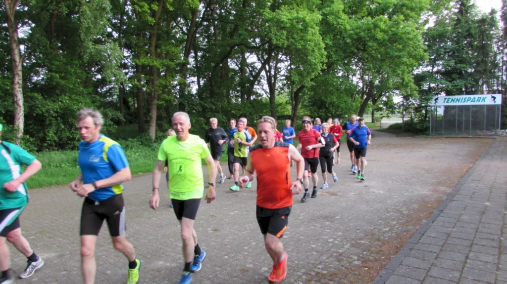 Gruppe von Laufenden in Sportbekleidung, die auf einem Weg im Tennispark joggen, umgeben von grüner Natur und Bäumen.
