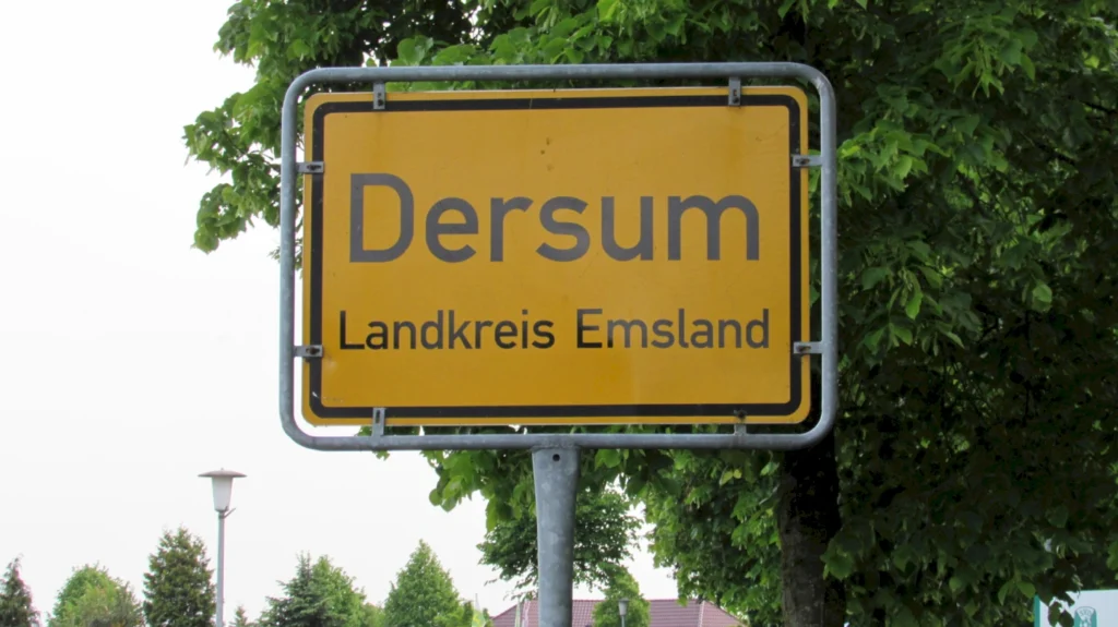 Ortsschild von Dersum im Landkreis Emsland, umgeben von Bäumen, unter bewölktem Himmel.