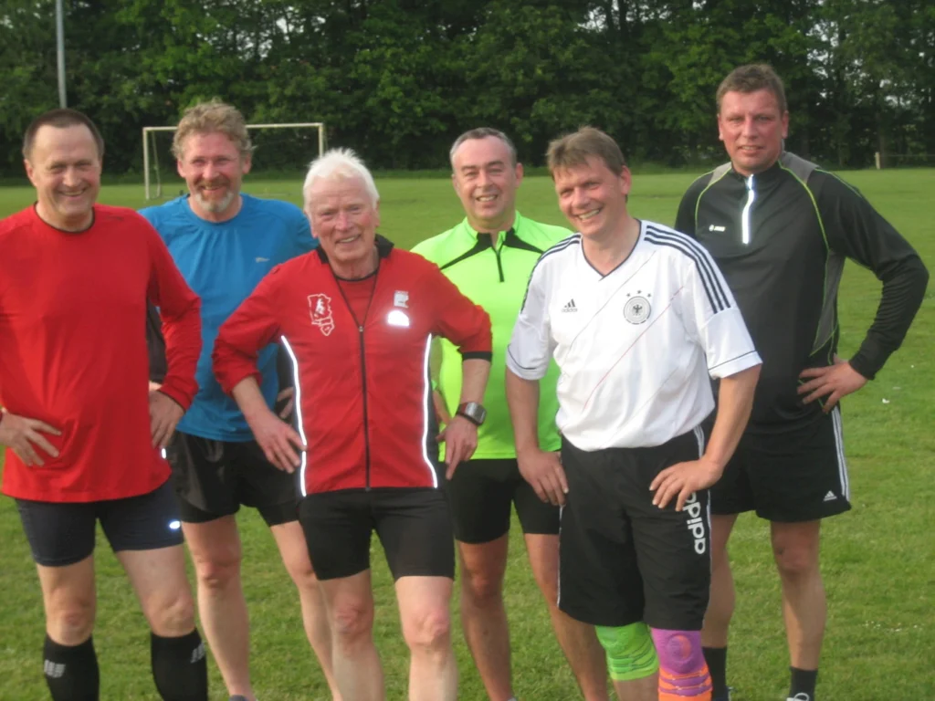 Gruppe von Männern beim Fußballtraining auf einem grünen Sportplatz, lächelnd in Sportkleidung versammelt.