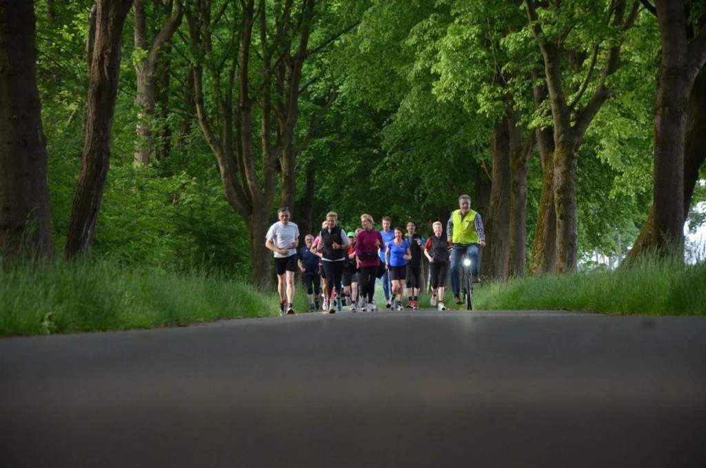 Eine Gruppe von Läufern joggt auf einem schmalen Weg, umgeben von Bäumen und grünem Gras, während ein Radfahrer sie begleitet. Ideal für Fitness und Outdoor-Aktivitäten.