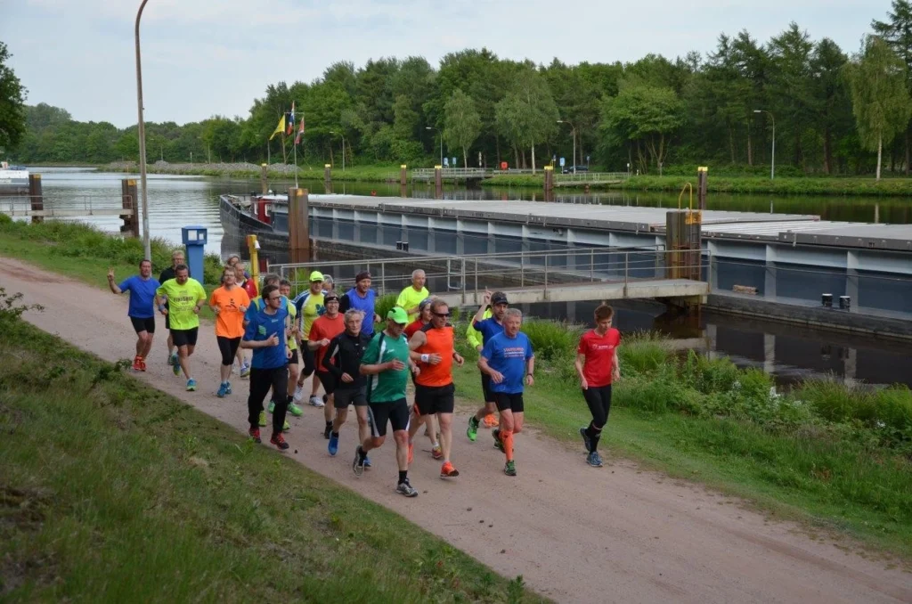 Gruppe von Läufern joggt entlang eines Kanals neben einer Schleuse, umgeben von grüner Natur und Bäumen.