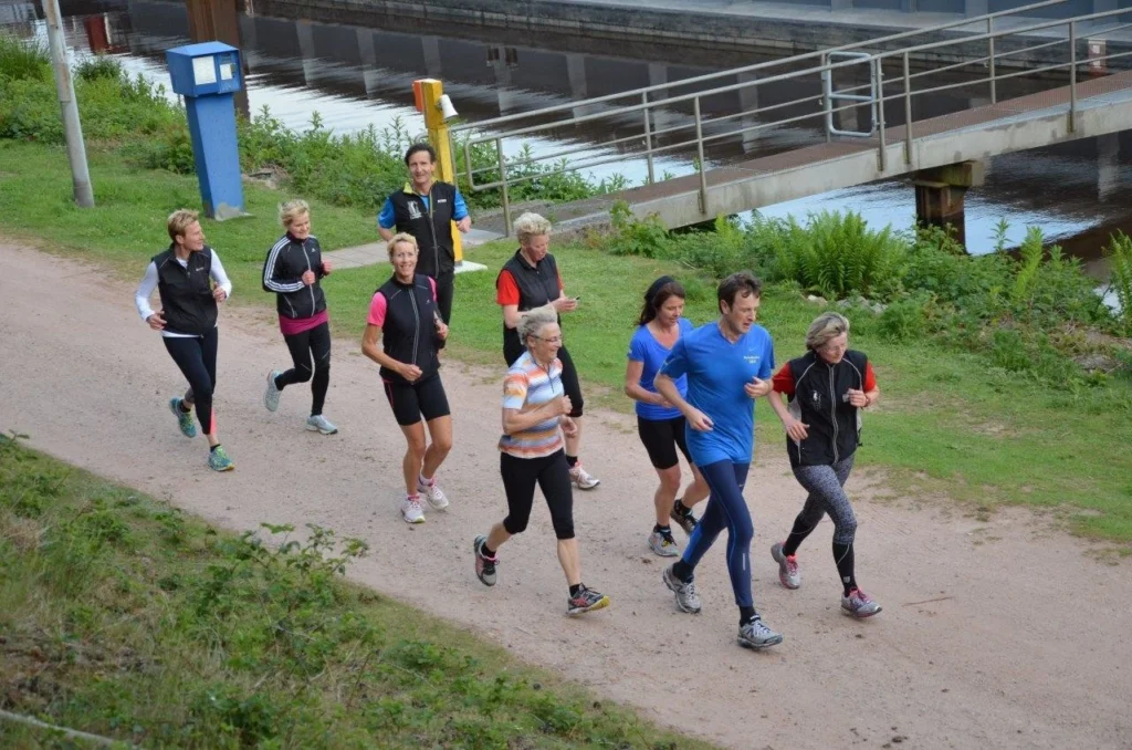 Gruppe von Menschen joggt gemeinsam auf einem Pfad entlang eines Flusses, gesäumt von Gras und Pflanzen.