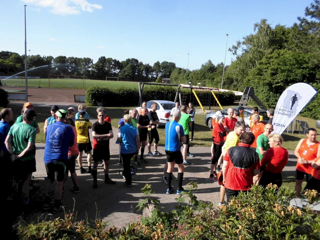 Gruppierung von Läufern in Sportkleidung bei einem Lauf-Event, umgeben von einem Sportplatz und Spielgeräten, mit einem Banner im Hintergrund, das die Veranstaltung präsentiert.