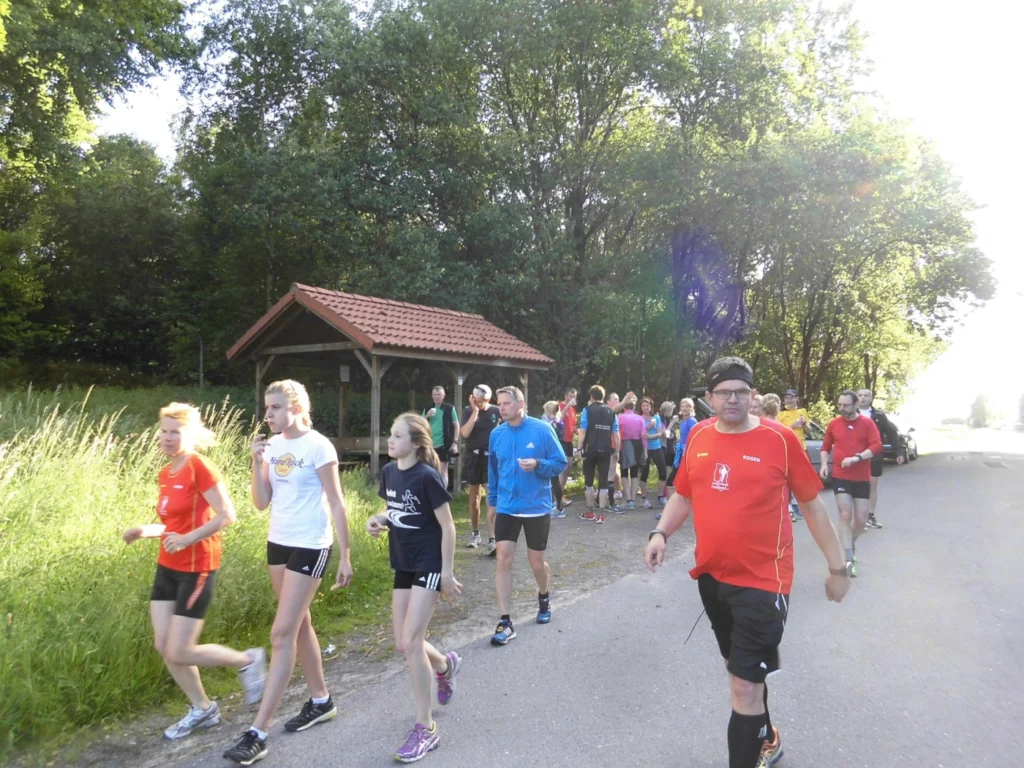 Gruppe von Menschen bei einem Lauf im Grünen an einem sonnigen Tag, umgeben von Bäumen und einem Pavillon im Hintergrund.
