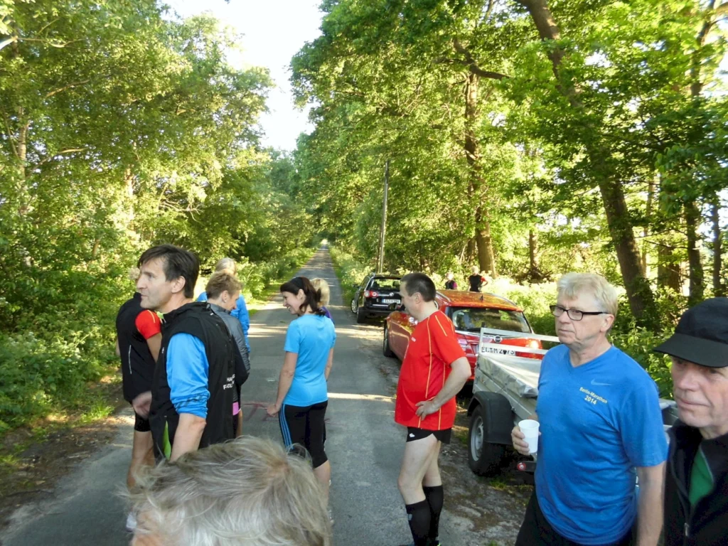 Läufer in blauen T-Shirts stehen auf einem schmalen Weg umgeben von Bäumen, während sie sich auf einen Wettkampf vorbereiten. Im Hintergrund sind Autos geparkt, und einige Teilnehmer unterhalten sich.