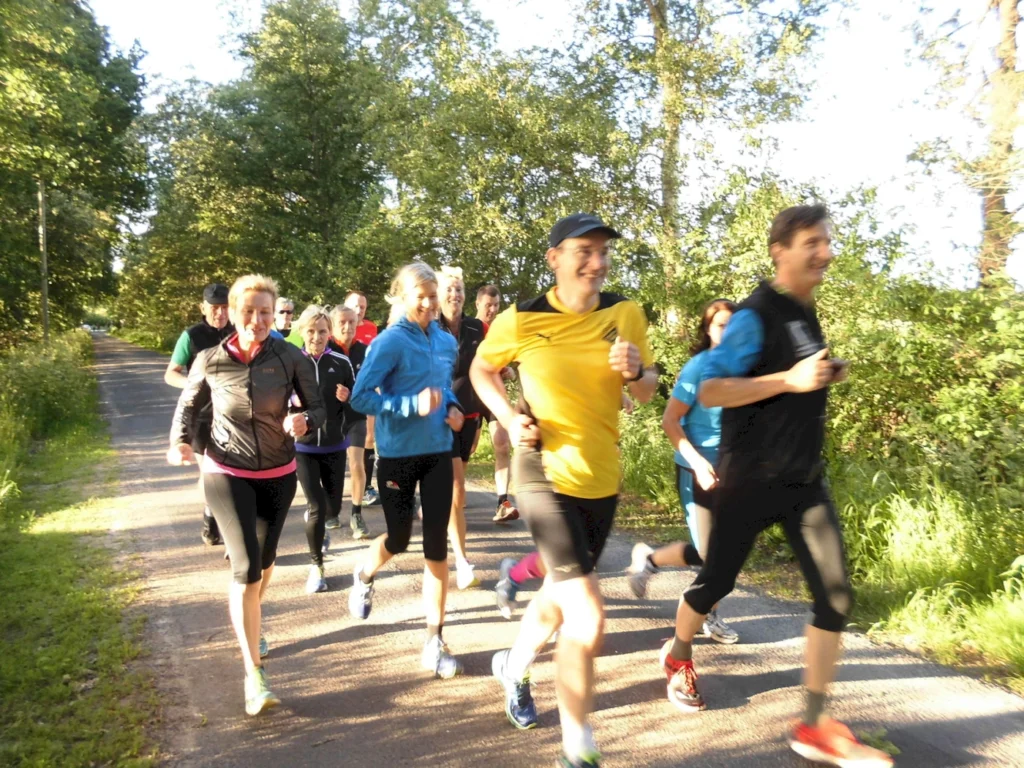 Gruppe von Menschen joggt auf einem Waldweg bei Sonnenschein, umgeben von grünen Bäumen, für Fitness und Spaß.