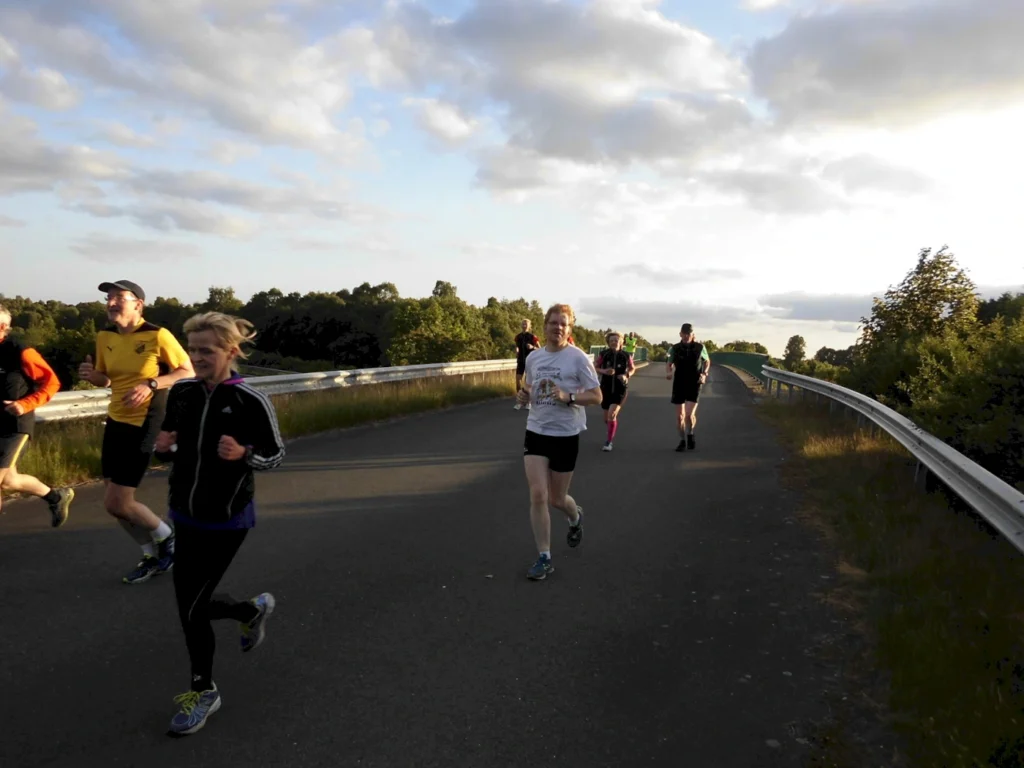 Gruppe von Läufern, die auf einer Straße mit grüner Landschaft im Hintergrund joggen, bei sonnigem Wetter und leicht bewölktem Himmel.