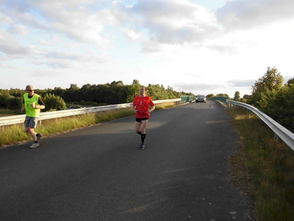 Zwei Jogger laufen bei bewölktem Himmel auf einer Straße. Ein Auto fährt im Hintergrund.