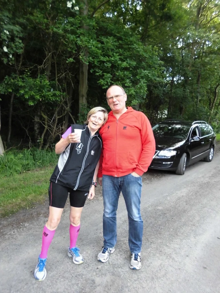 Zwei Personen in Sportkleidung lächelnd auf einem Waldweg mit Auto im Hintergrund.
