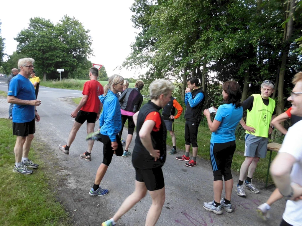 Gruppe von Läufern in Sportkleidung, die an einer Straßenecke stehen und sich aufwärmen. Die Szene zeigt eine Mischung aus Männern und Frauen, die in einer natürlichen Umgebung mit Bäumen und einer Straße im Hintergrund aktiv sind.