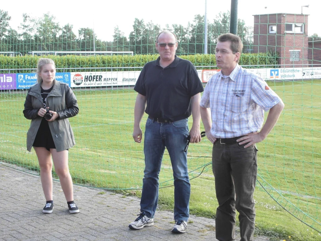 Personen stehen auf einem Sportplatz neben einem Fußballfeld, eine Frau hält eine Kamera.
