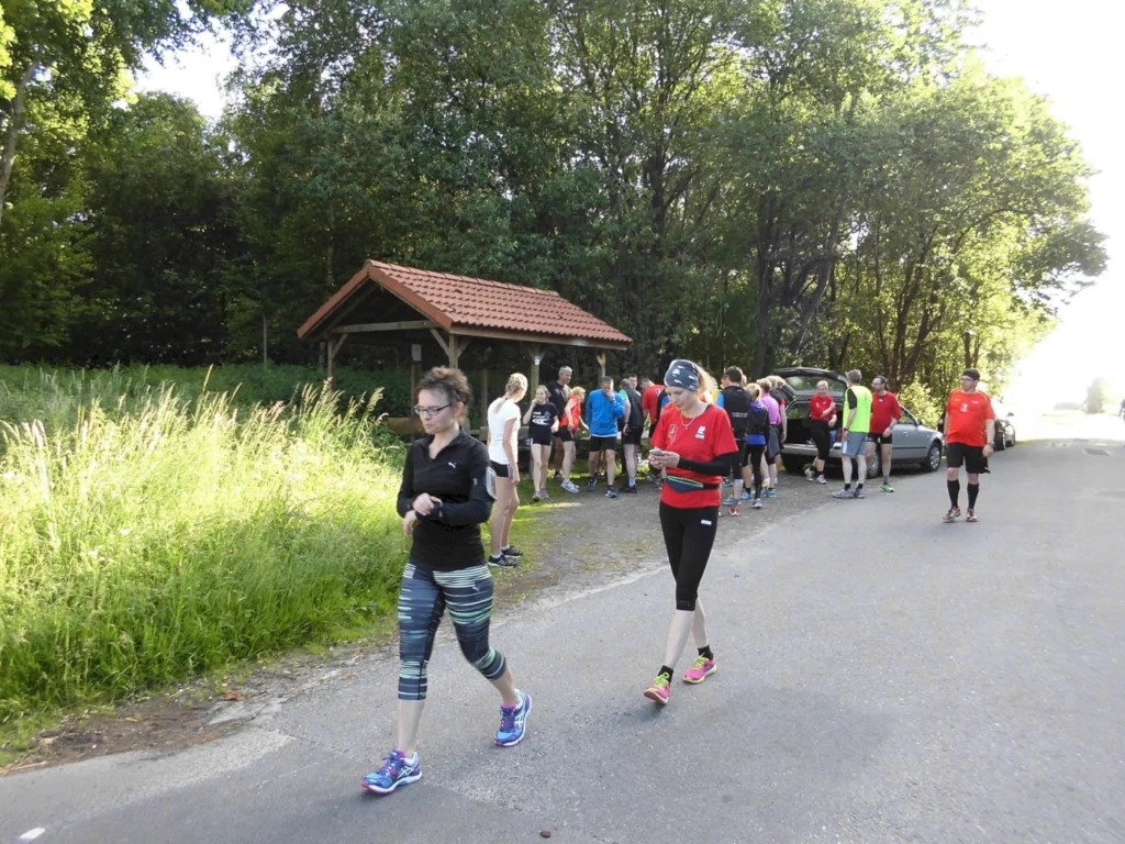 Gruppe von Läufern, die sich an einem sonnigen Morgen versammelt, um an einem Lauf teilzunehmen. Im Hintergrund sind Bäume und eine Überdachung zu sehen, während einige Teilnehmer ihre Uhren überprüfen und sich aufwärmen.
