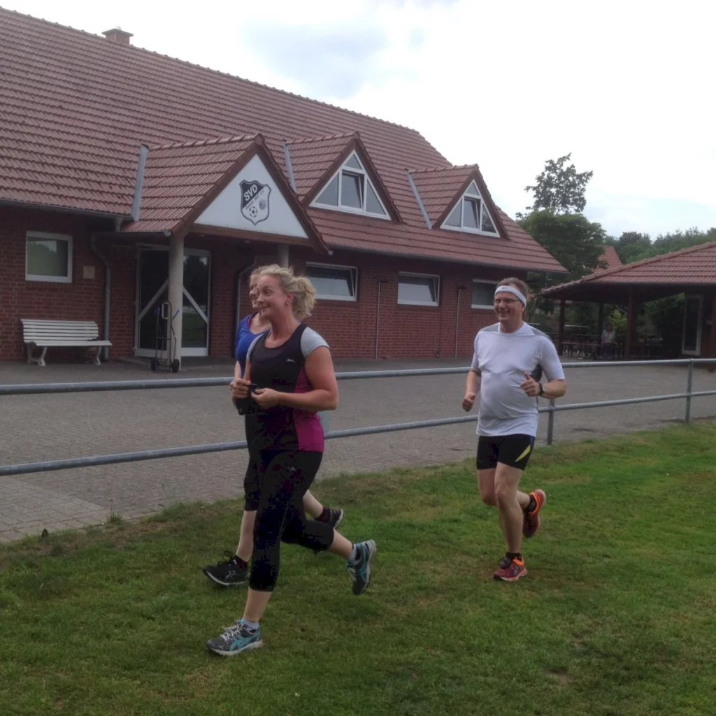 Drei Personen joggen fröhlich auf einem Grasfeld vor einem Vereinsgebäude mit rotem Ziegeldach. Im Hintergrund sind Sitzgelegenheiten und Bäume sichtbar.