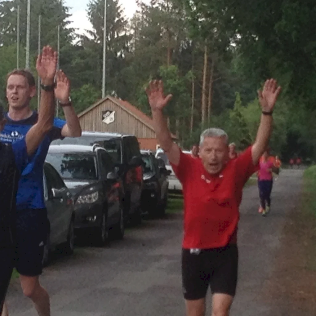Mehrere Personen joggen mit erhobenen Armen auf einer Waldstraße, Autos im Hintergrund.