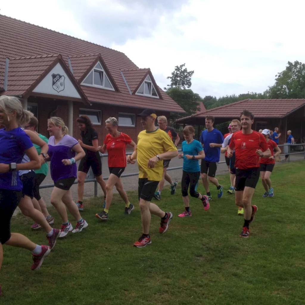 Gruppe von Läufern in sportlicher Kleidung, die auf einer Wiese vor einem Vereinsgebäude joggen. Die Teilnehmer sind in verschiedenen Farben gekleidet, darunter Rot, Blau und Gelb. Im Hintergrund sind Bäume und ein weiteres Gebäude zu sehen, was eine freundliche Sportatmosphäre vermittelt.