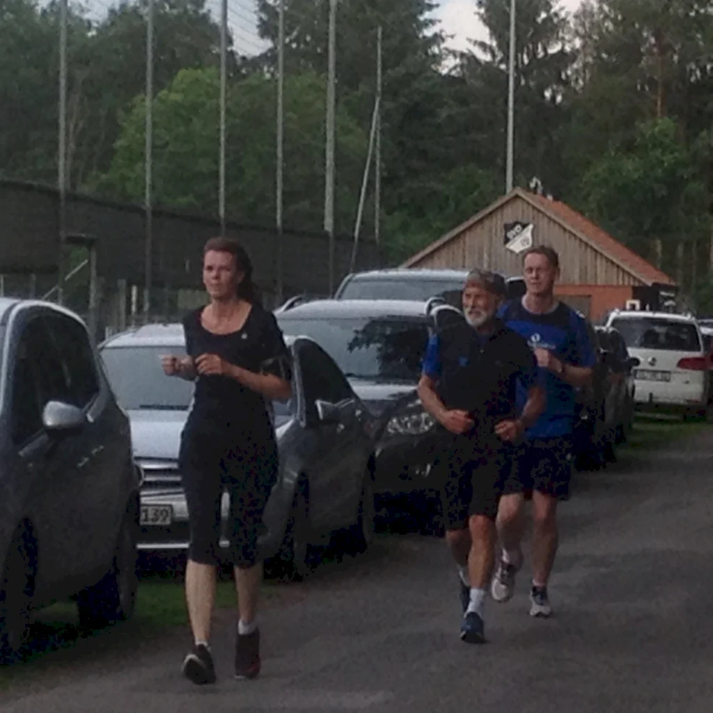 Drei Personen joggen auf einem Weg zwischen parkenden Autos, umgeben von Bäumen und einem Holzgebäude im Hintergrund.