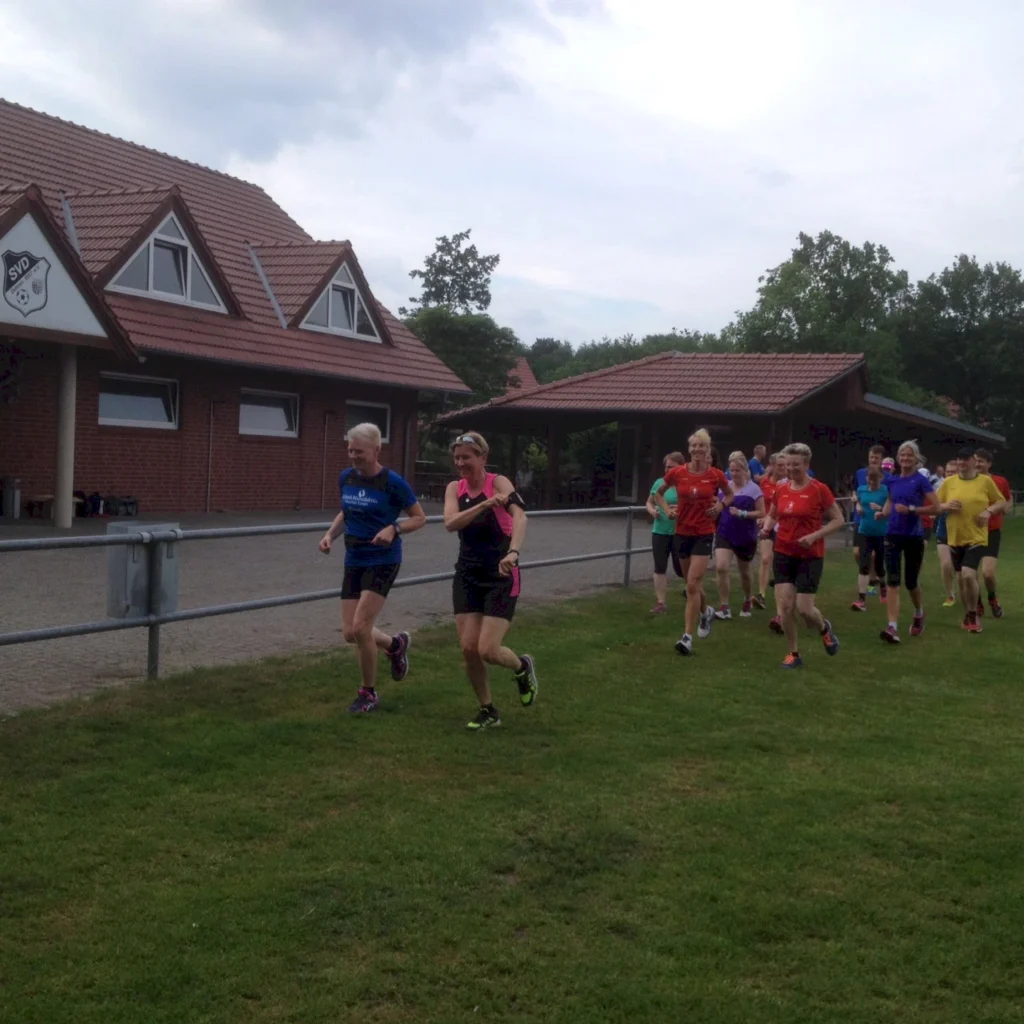 Gruppe von Läufern in Sportkleidung, die auf einem Rasen vor einem Vereinsgebäude joggen. Die Teilnehmer sind männlich und weiblich, in verschiedenen Farben gekleidet, und zeigen eine aktive Laufbewegung.