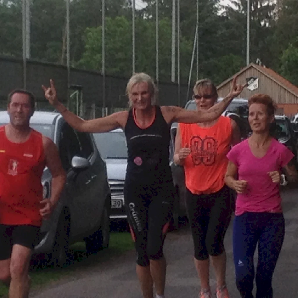 Vier Menschen joggen auf einer Straße neben parkenden Autos, eine Person streckt die Arme in die Luft.