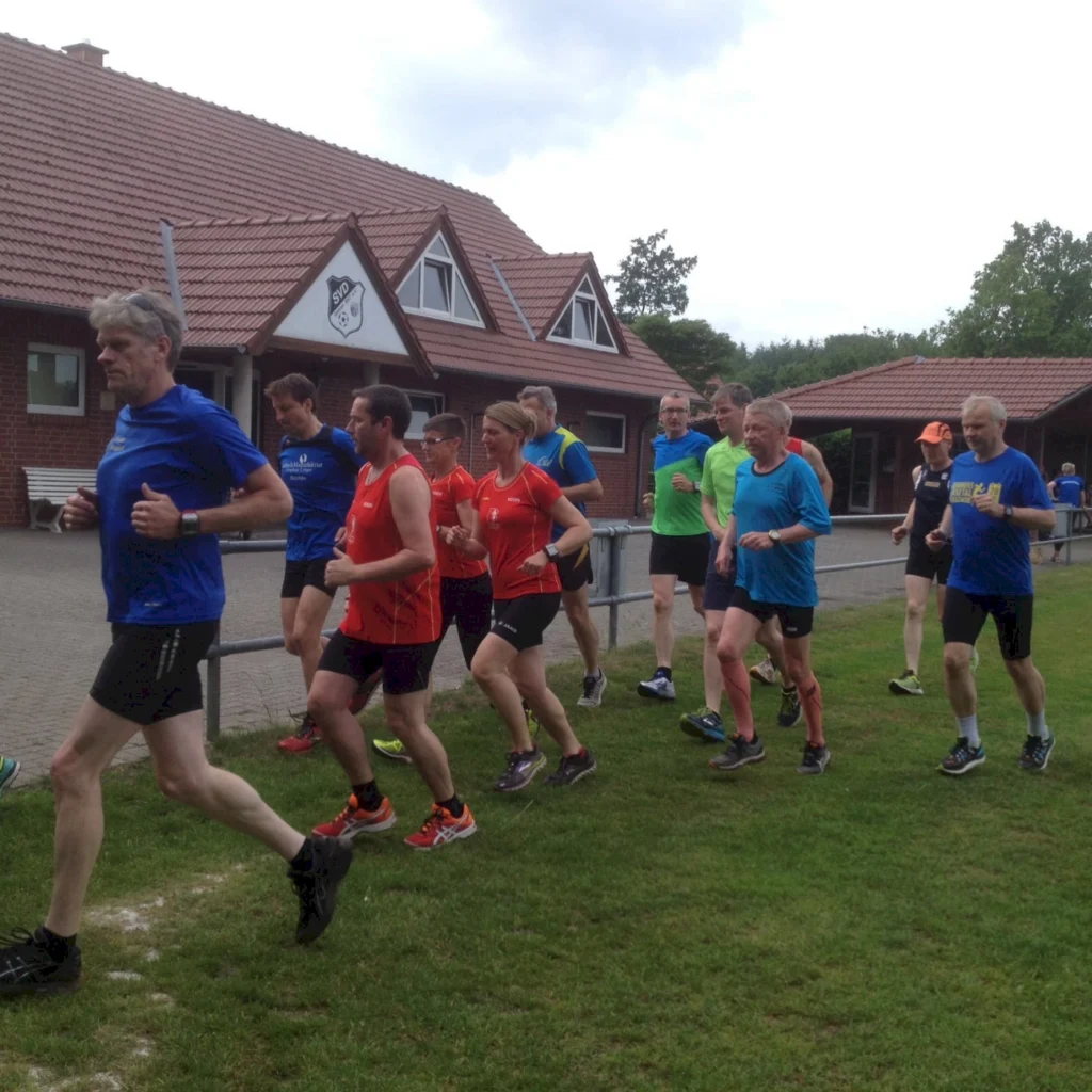 Gruppe von Menschen joggt auf einer Wiese vor einem Gebäude. Sie tragen bunte Sportkleidung.