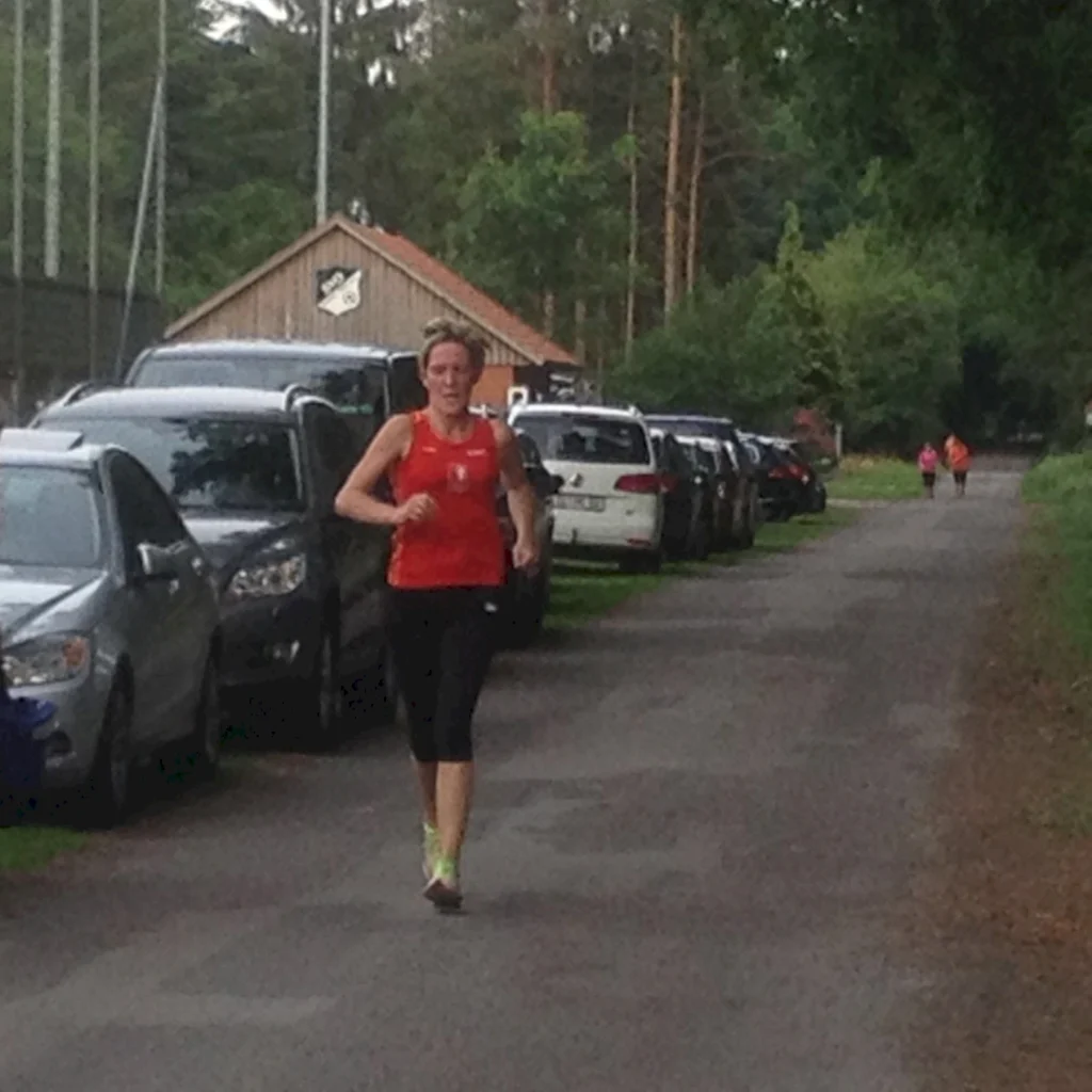 Frau joggt auf einer ländlichen Straße mit geparkten Autos und einem kleinen Gebäude im Hintergrund.