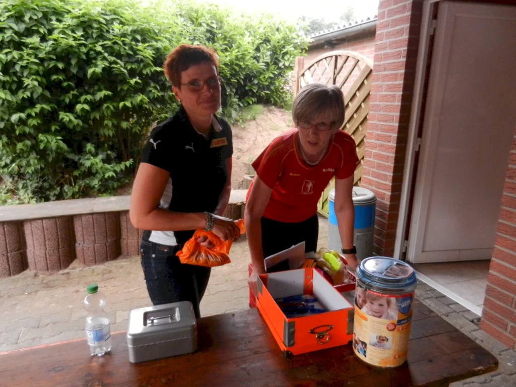 Zwei Frauen bei der Organisation einer Tombola, draußen neben einem Tisch mit Spendenboxen und einer Metalldose.