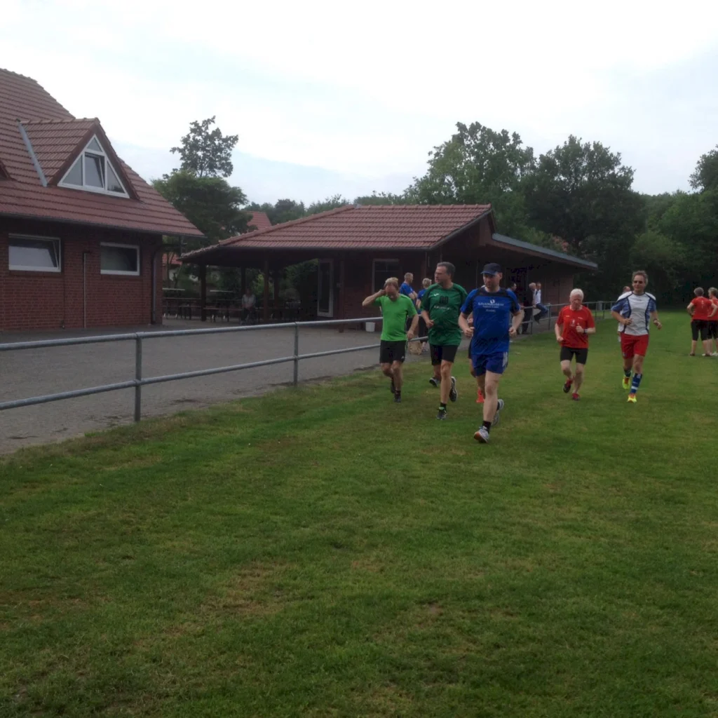Gruppentraining von Läufern auf einem Rasenplatz vor einem Vereinsgebäude, umgeben von Bäumen, an einem bewölkten Tag.