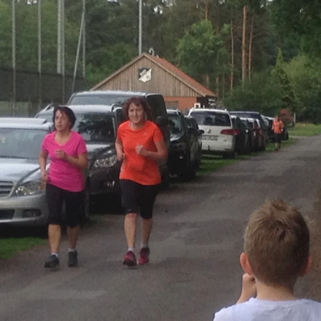 Zwei Frauen joggen auf einem Waldweg, während ein Kind am Rand zuschaut. Autos parken seitlich entlang des Weges.