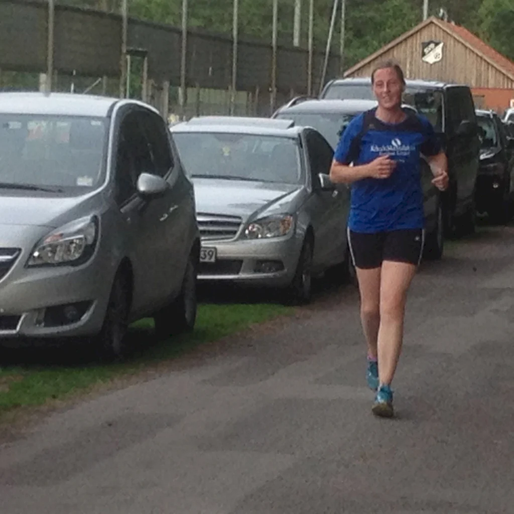 Eine lächelnde Frau in Sportkleidung joggt auf einem Weg entlang geparkter Autos, umgeben von einer grünen Landschaft.