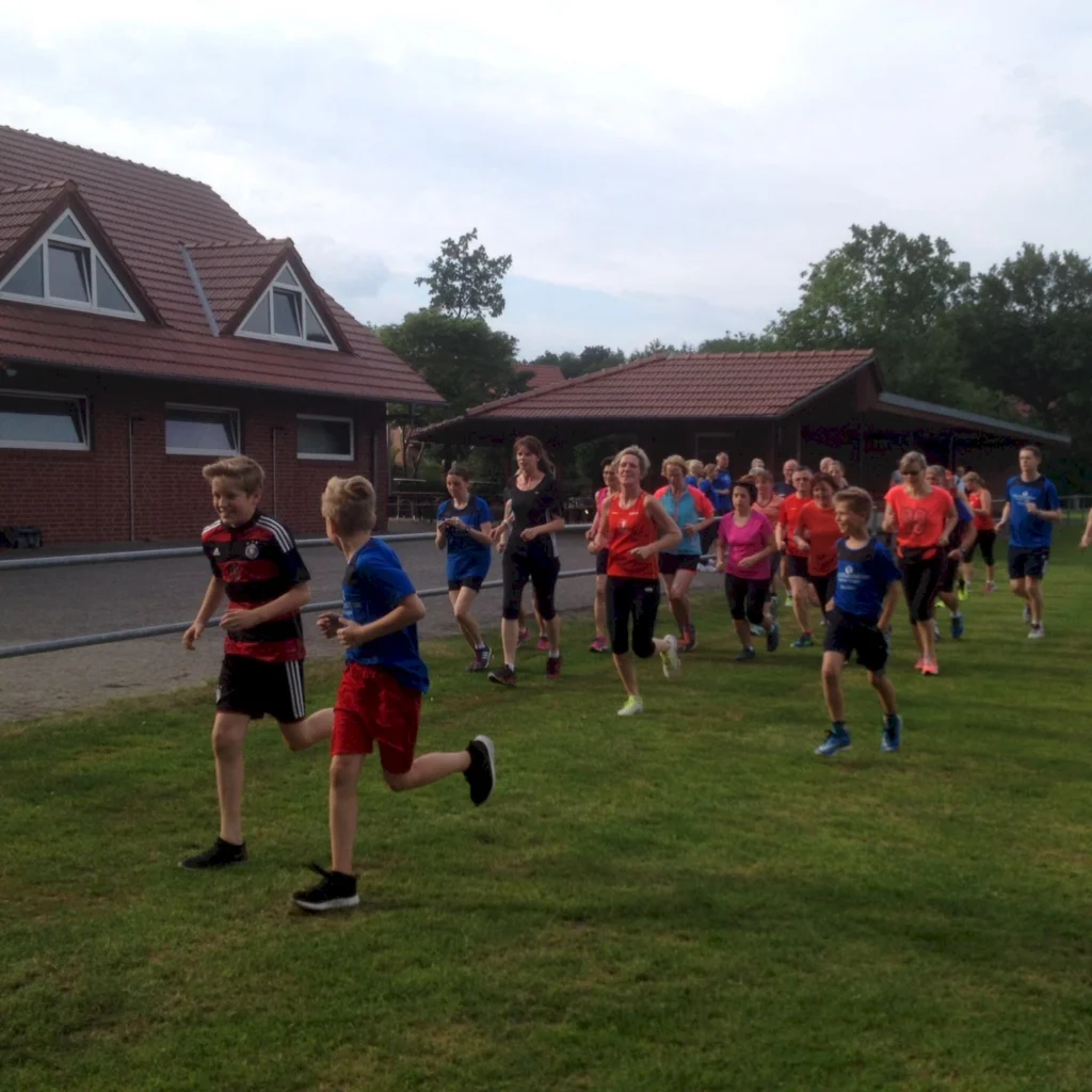 Gruppe von Menschen joggt auf einem grünen Feld vor Backsteingebäuden, sonniger Tag, Freizeitaktivität im Freien.