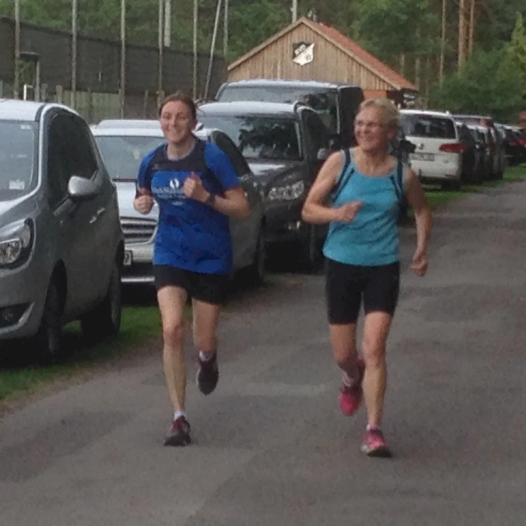 Zwei Frauen joggen fröhlich auf einem Parkplatz mit Autos im Hintergrund.