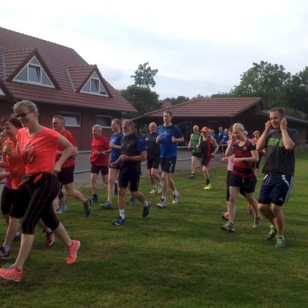 Gruppe von Läufern in Sportkleidung, die auf einer Wiese joggen, umgeben von Wohngebäuden und Bäumen.